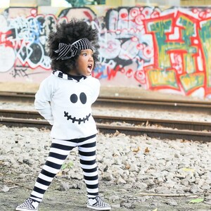 May include: A young child wearing a white hoodie with a black ghost face design and black and white striped pants. The child is standing in front of a graffiti wall.