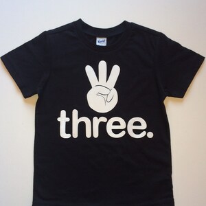 Może przedstawiać: Czarny T-shirt z białym nadrukiem dłoni pokazującej znak liczby trzy i słowem "three".