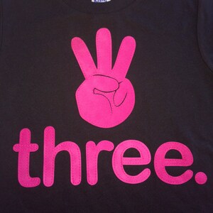 Może przedstawiać: Czarny T-shirt z r&oacute;żową cyfrą trzy i słowem "three" w kolorze r&oacute;żowym.