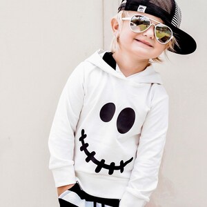 May include: A white hoodie with a black stitched-smile face design. The hoodie has a black hood and is worn by a child wearing black and white striped pants and a black mesh baseball cap with a white logo.