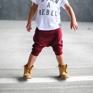 以下が含まれることがあります： 白いTシャツに黒い文字で「Born a Rebel」と書かれたものを着ている若い男の子。彼は、ワインレッドのキュロットと茶色のブーツを履いています。