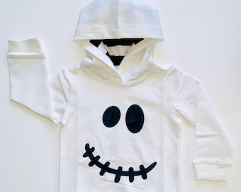 May include: A white long-sleeve hooded sweatshirt featuring a black graphic of two oval eyes and a stitched smile on the front. The interior of the hood is lined with a black and white horizontal striped fabric, and the sleeves have ribbed cuffs.
