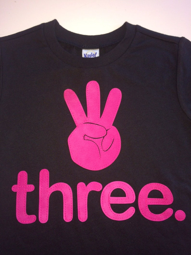 Może przedstawiać: Czarny T-shirt z r&oacute;żową cyfrą trzy i słowem "three" w kolorze r&oacute;żowym.