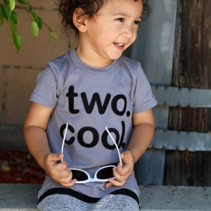 Puede incluir: Un ni&ntilde;o peque&ntilde;o con una camiseta gris con el texto "two. cool." impreso en negro. El ni&ntilde;o lleva pantalones grises y negros a rayas y sostiene unas gafas de sol blancas.