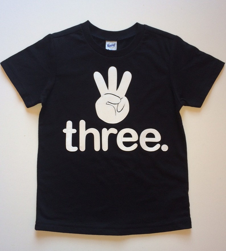 Może przedstawiać: Czarny T-shirt z białym nadrukiem dłoni pokazującej znak liczby trzy i słowem "three".