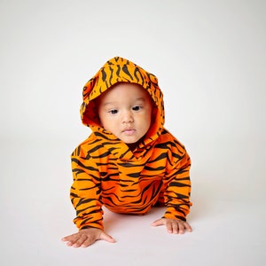 May include: A baby wearing an orange and black tiger-striped hooded onesie. The baby is crawling on a white surface.