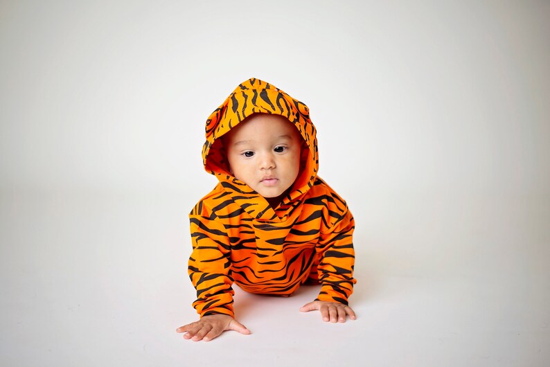 May include: A baby wearing an orange and black tiger-striped hooded onesie. The baby is crawling on a white surface.
