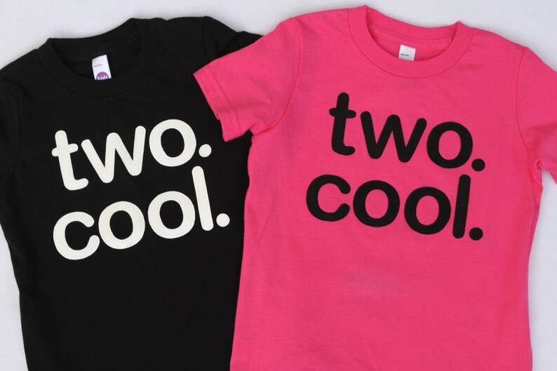 Puede incluir: Dos camisetas, una negra y otra rosa, con el texto "two. cool." impreso en ellas. La camiseta negra tiene texto blanco y la camiseta rosa tiene texto negro.