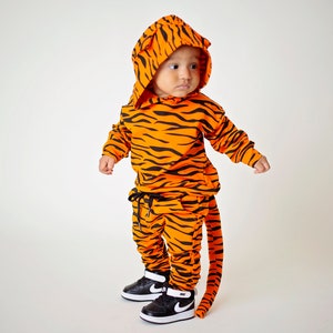 May include: A child wearing a tiger-print hooded sweatshirt and matching pants with a tail. The outfit is orange and black with a black drawstring on the pants. The child is wearing black and white sneakers.