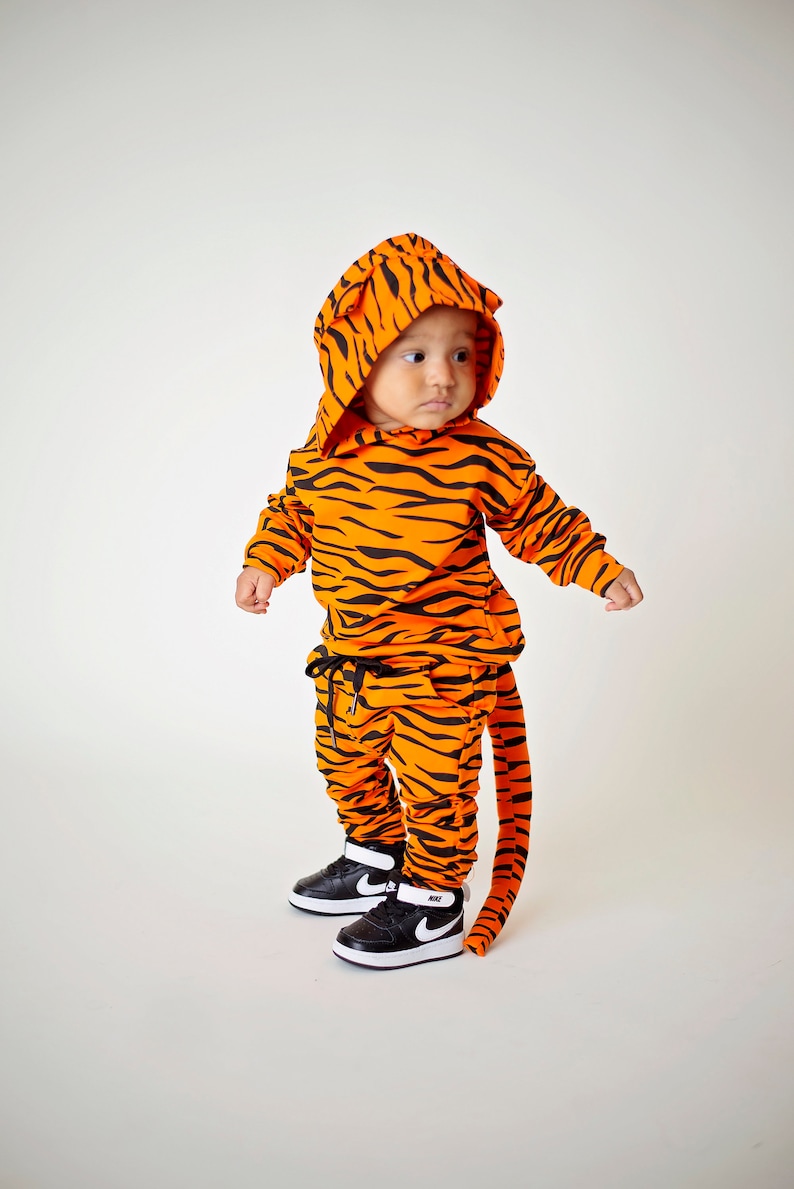 May include: A child wearing a tiger-print hooded sweatshirt and matching pants with a tail. The outfit is orange and black with a black drawstring on the pants. The child is wearing black and white sneakers.