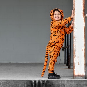 May include: A child wearing an orange and black tiger costume with a tail. The child is smiling and holding onto a white post.