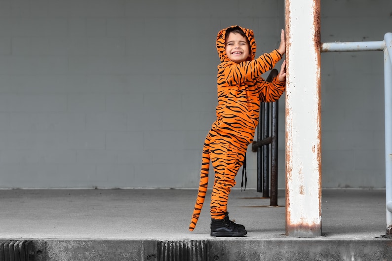 May include: A child wearing an orange and black tiger costume with a tail. The child is smiling and holding onto a white post.