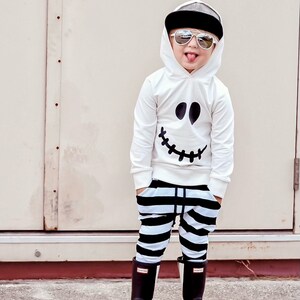 May include: A young child wearing a white hoodie with a black ghost face design, black and white striped pants, and black skeleton-patterned rain boots. The child is wearing sunglasses and has a black baseball cap on their head.