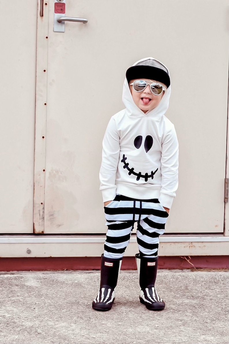 May include: A young child wearing a white hoodie with a black ghost face design, black and white striped pants, and black skeleton-patterned rain boots. The child is wearing sunglasses and has a black baseball cap on their head.