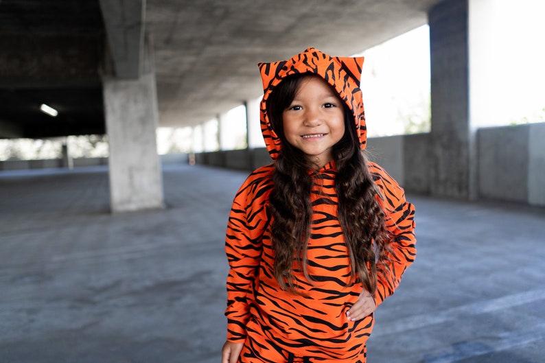 May include: A young person wearing an orange and black tiger print hoodie with a hood pulled up. The hoodie has ears on the top. The person is smiling and looking at the camera.