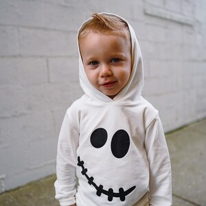 May include: A young child wearing a white hooded sweatshirt with a black ghost face design. The child is looking at the camera and smiling.