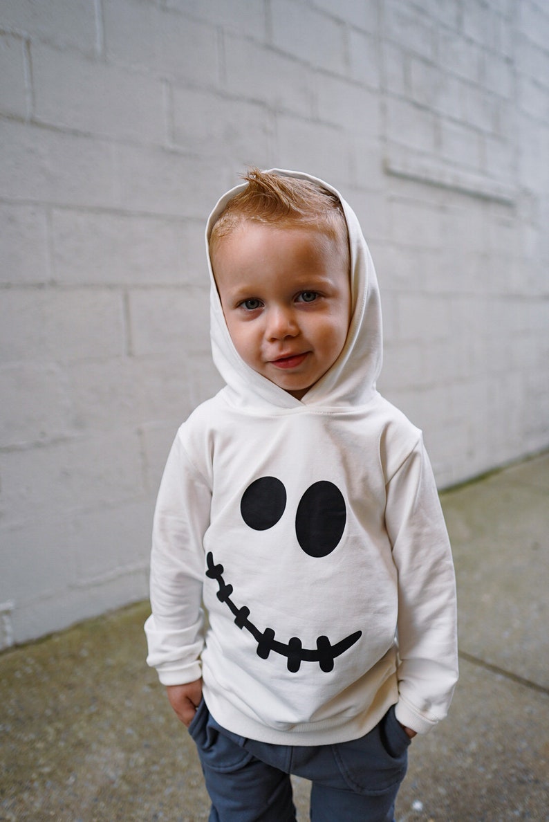 May include: A young child wearing a white hooded sweatshirt with a black ghost face design. The child is looking at the camera and smiling.