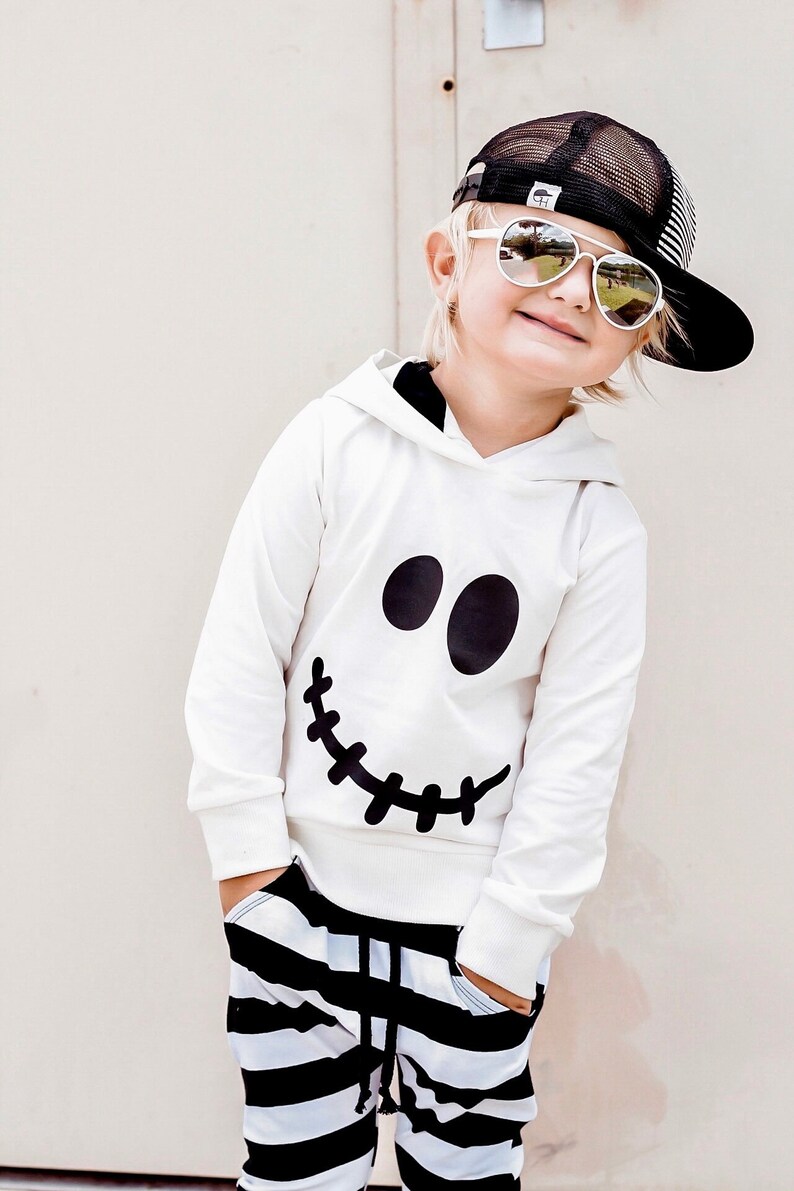 May include: A white hoodie with a black stitched-smile face design. The hoodie has a black hood and is worn by a child wearing black and white striped pants and a black mesh baseball cap with a white logo.