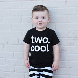 Puede incluir: Un ni&ntilde;o peque&ntilde;o con una camiseta negra con el texto "two. cool." en letras blancas. Tambi&eacute;n lleva pantalones cortos a rayas blancas y negras.