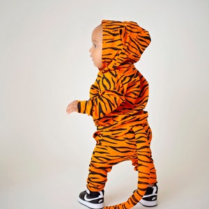 May include: A child wearing an orange and black tiger costume with a tail. The costume has a hood and the child is wearing black sneakers.