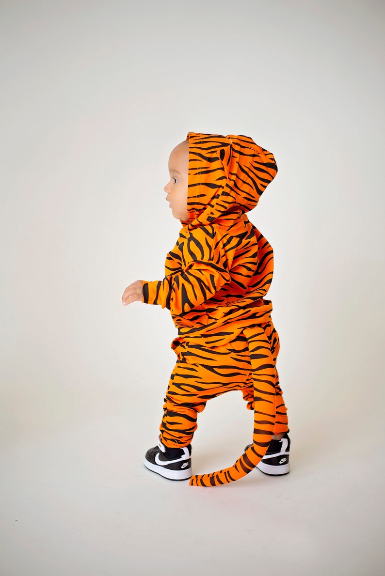 May include: A child wearing an orange and black tiger costume with a tail. The costume has a hood and the child is wearing black sneakers.