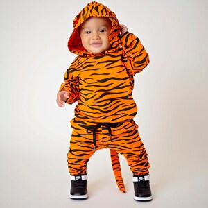 May include: A baby wearing a tiger-print hooded jumpsuit with a tail. The jumpsuit is orange and black with a black drawstring waist. The baby is wearing black sneakers with white accents.