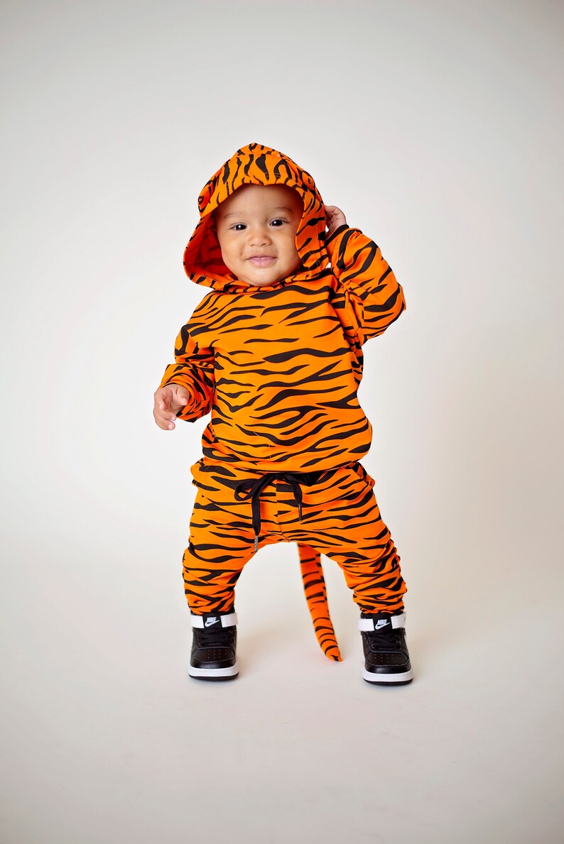 May include: A baby wearing a tiger-print hooded jumpsuit with a tail. The jumpsuit is orange and black with a black drawstring waist. The baby is wearing black sneakers with white accents.