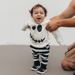 May include: A baby wearing a white long-sleeved shirt with a black and white ghost face design and black and white striped pants. The baby is smiling and looking at the camera.