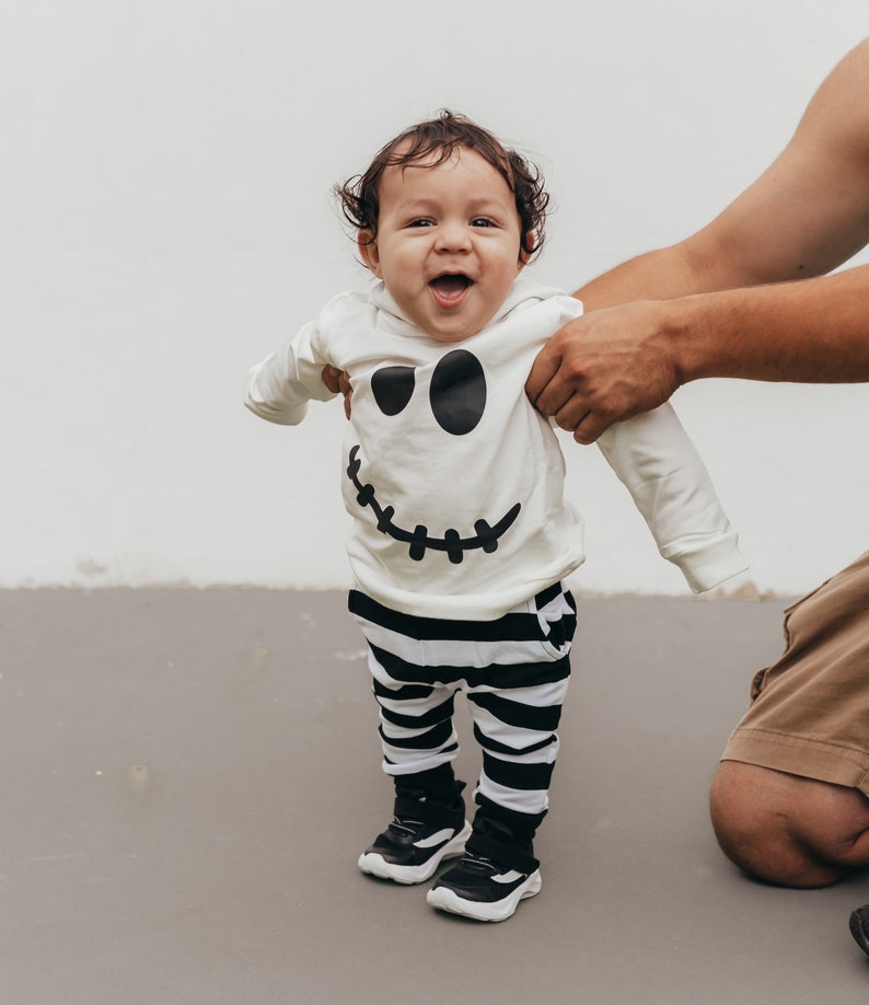 May include: A baby wearing a white long-sleeved shirt with a black and white ghost face design and black and white striped pants. The baby is smiling and looking at the camera.