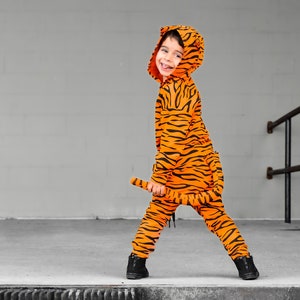 May include: A child wearing an orange and black tiger costume with a tail. The child is standing on a concrete surface with a metal railing in the background.