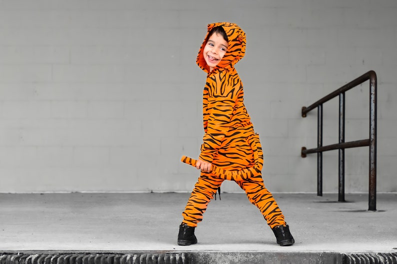 May include: A child wearing an orange and black tiger costume with a tail. The child is standing on a concrete surface with a metal railing in the background.