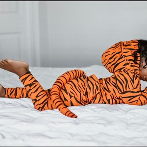 May include: A child wearing a tiger-print hooded jumpsuit with a tail. The jumpsuit is orange and black with a striped pattern. The child is lying on a white bed.