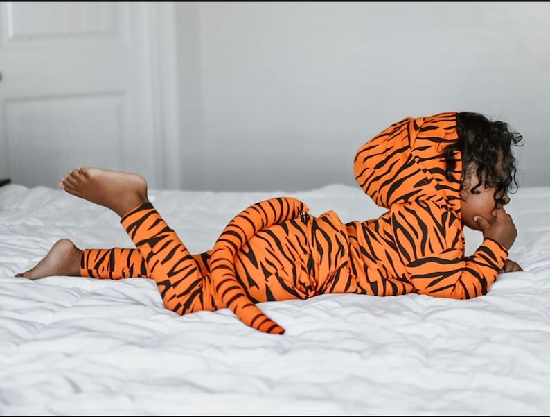 May include: A child wearing a tiger-print hooded jumpsuit with a tail. The jumpsuit is orange and black with a striped pattern. The child is lying on a white bed.