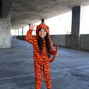 May include: A young person dressed in an orange and black tiger costume with a tail. They are smiling and holding their hand up in a claw-like gesture.