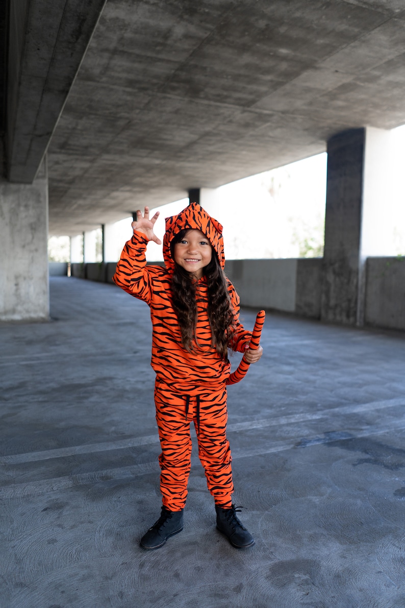 May include: A young person dressed in an orange and black tiger costume with a tail. They are smiling and holding their hand up in a claw-like gesture.