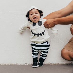 May include: A baby wearing a white hooded costume with a black ghost face design and black and white striped pants. The baby is standing in front of a white wall.