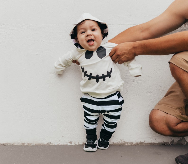 May include: A baby wearing a white hooded costume with a black ghost face design and black and white striped pants. The baby is standing in front of a white wall.