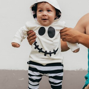 May include: A baby wearing a white hooded onesie with a black ghost face design and black and white striped pants. The baby is standing and looking up.