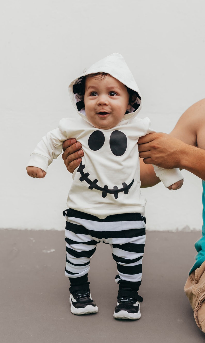 May include: A baby wearing a white hooded onesie with a black ghost face design and black and white striped pants. The baby is standing and looking up.