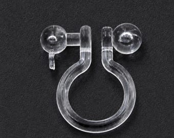 May include: A transparent plastic earring clip, featuring a circular loop and two spherical ends. The clip has a small hook on one side, designed for earlobe attachment. Set against a plain black backdrop.