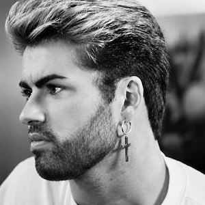 Op de afbeelding: Zwart-wit portret van een persoon met een zilveren oorring met een kruisvormige hanger. De persoon heeft gestyled haar en een baard. De oorring is het centrale element.