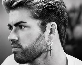 May include: Black and white portrait of a person wearing a silver hoop earring with a cross pendant. The person has short, styled hair and a beard. The earring is the focal point.