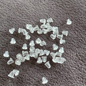 Könnte beinhalten: Eine Ansammlung klarer, durchscheinender Ohrringverschlüsse auf einer strukturierten, dunkelgrauen Oberfläche. Die Ohrringverschlüsse sind klein, mit einem glockenförmigen Design und scheinen aus weichem Kunststoff oder Silikon zu bestehen.