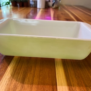 May include: A white ceramic loaf pan with a slightly off-white color. The pan is sitting on a wooden surface.