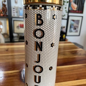 May include: A tall, cylindrical travel mug with a gold-colored lid and rim. The body features a white honeycomb pattern with black accents and the word "BONJOUR" in gold lettering. Small black and gold floral designs are scattered across the surface.