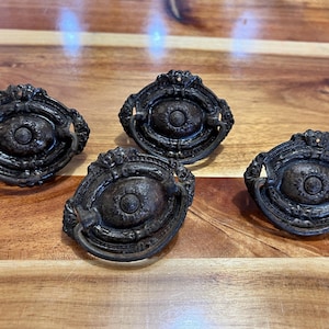 Op de afbeelding: Vier antieke bronzen lade-knoppen met een ovaal ontwerp en een centraal cirkelvormig detail. De knoppen hebben sierlijke details en een ringhandvat. Ze zijn gerangschikt op een houten oppervlak.