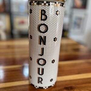 May include: A tall, cylindrical tumbler with a gold rim and lid. The tumbler features a white honeycomb pattern with the word "BONJOUR" in gold lettering. Small black floral accents are scattered around the design.