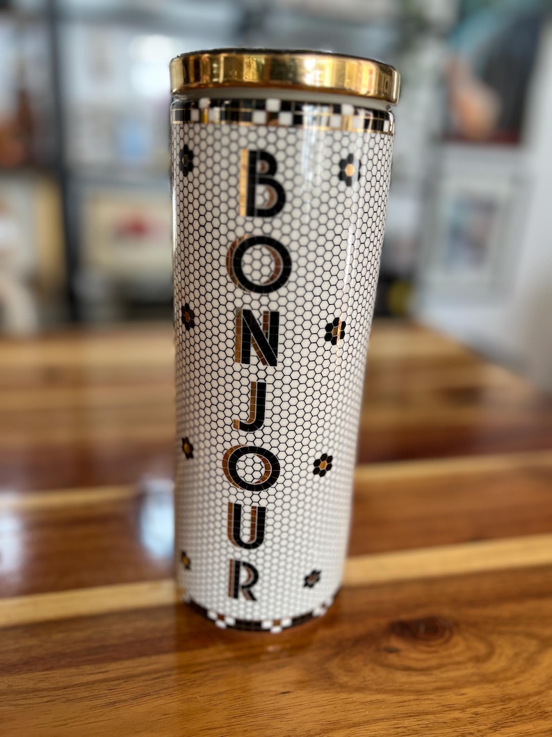 May include: A tall, cylindrical tumbler with a gold rim and lid. The tumbler features a white honeycomb pattern with the word "BONJOUR" in gold lettering. Small black floral accents are scattered around the design.
