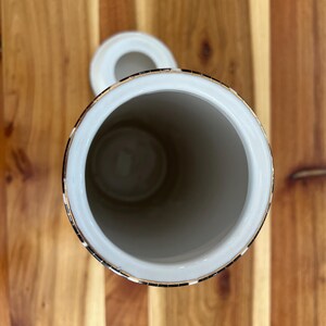 May include: A white ceramic jar with a lid, featuring a gold rim and a decorative black and gold band. The jar is cylindrical with a wide opening, set against a wooden surface.