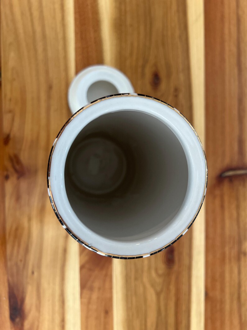 May include: A white ceramic jar with a lid, featuring a gold rim and a decorative black and gold band. The jar is cylindrical with a wide opening, set against a wooden surface.